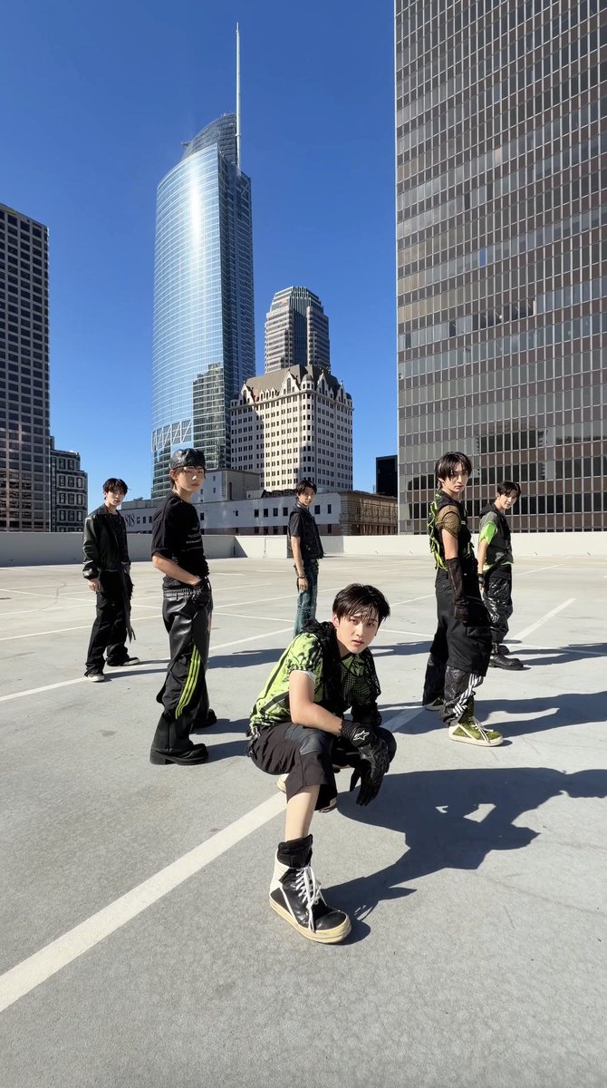 A group of six young men in black and green outfits pose on a rooftop parking lot with a city skyline and a glass tower in the background.