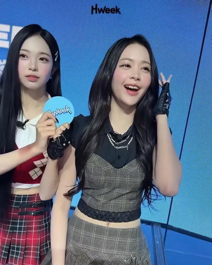 Two young women pose at what appears to be a "Hweek" event, with one holding a blue "Hweek" fan and making a peace sign while smiling brightly, while the other with long black hair stands beside her. The text "Hweek" is visible at the top of the image and on the fan, suggesting a Korean entertainment industry event, though the specific identity referenced as "carmen" in the caption isn't clearly verifiable from the image alone.