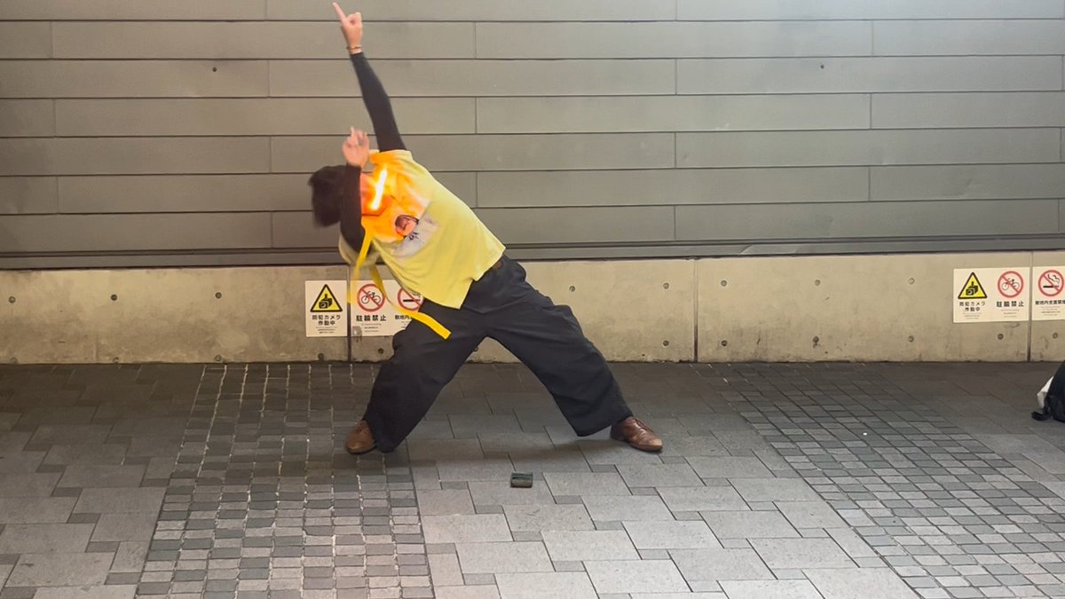 A person in a yellow vest and dark pants striking a dramatic action pose on a paved walkway near a concrete wall with warning signs.