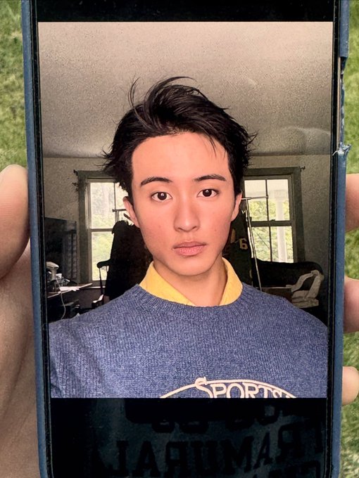 A young man with dark hair poses for a selfie displayed on a phone screen, wearing a gray sweater with a yellow collar and the word "SPORTS" visible.