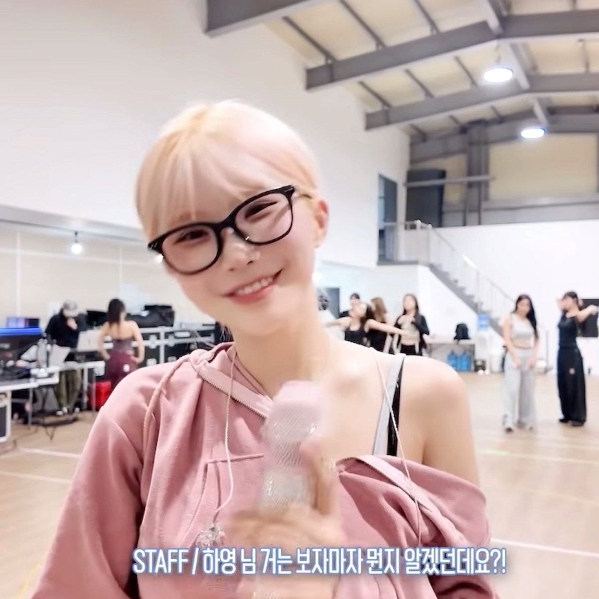 A close-up of a blonde-haired person wearing black glasses and a pink top in what appears to be a dance studio, with Korean text at the bottom reading 'STAFF / 하영 님 거는 보지마자 뭔지 알겠던데요?!'.