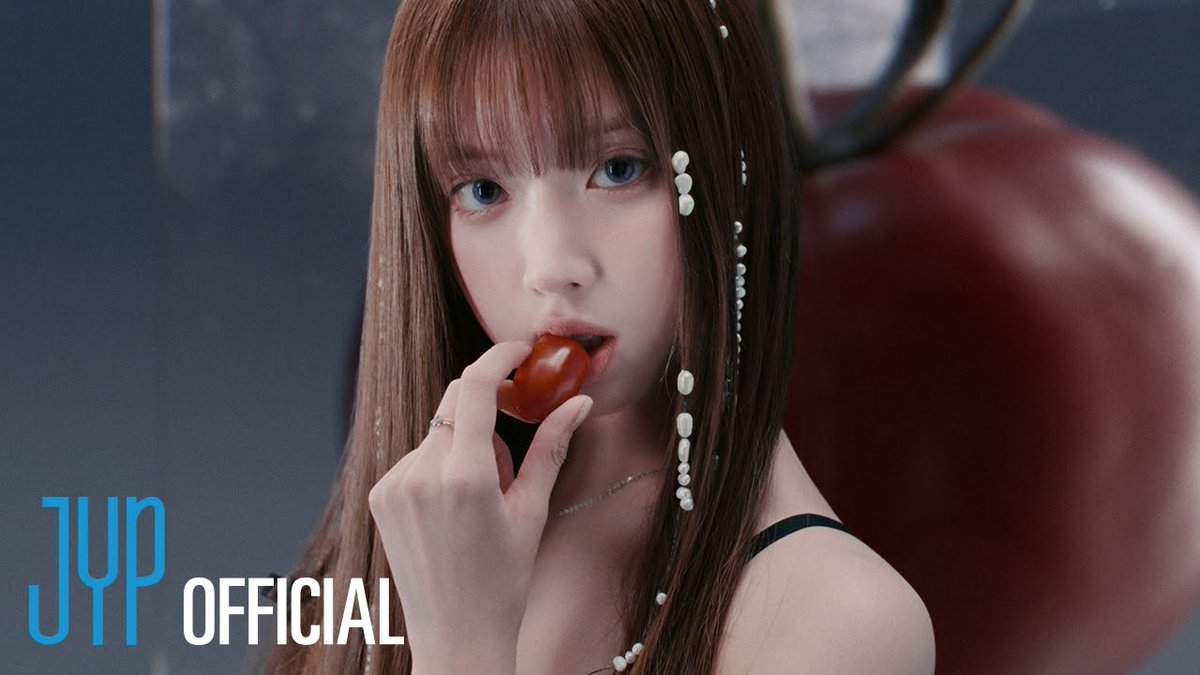 A still from a JYP OFFICIAL video shows a young woman with long brown hair and beaded hair accessories holding a small red tomato near her lips.