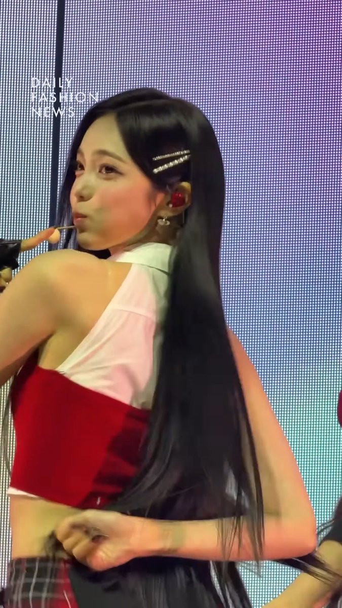 A young woman with long black hair and pearl hair clips wearing a white top and red corset, captured mid-performance with "DAILY FASHION NEWS" watermark visible.