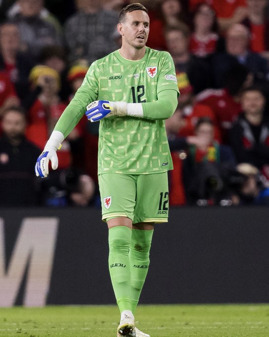Danny, wearing a green goalkeeper kit with the number 12 and "SUZUKI" sponsor logo, stands on the pitch during Cymru's victory over Norway last night. The image shows him in his Wales national team goalkeeper uniform, complete with gloves and matching green socks, against a backdrop of cheering fans in red.