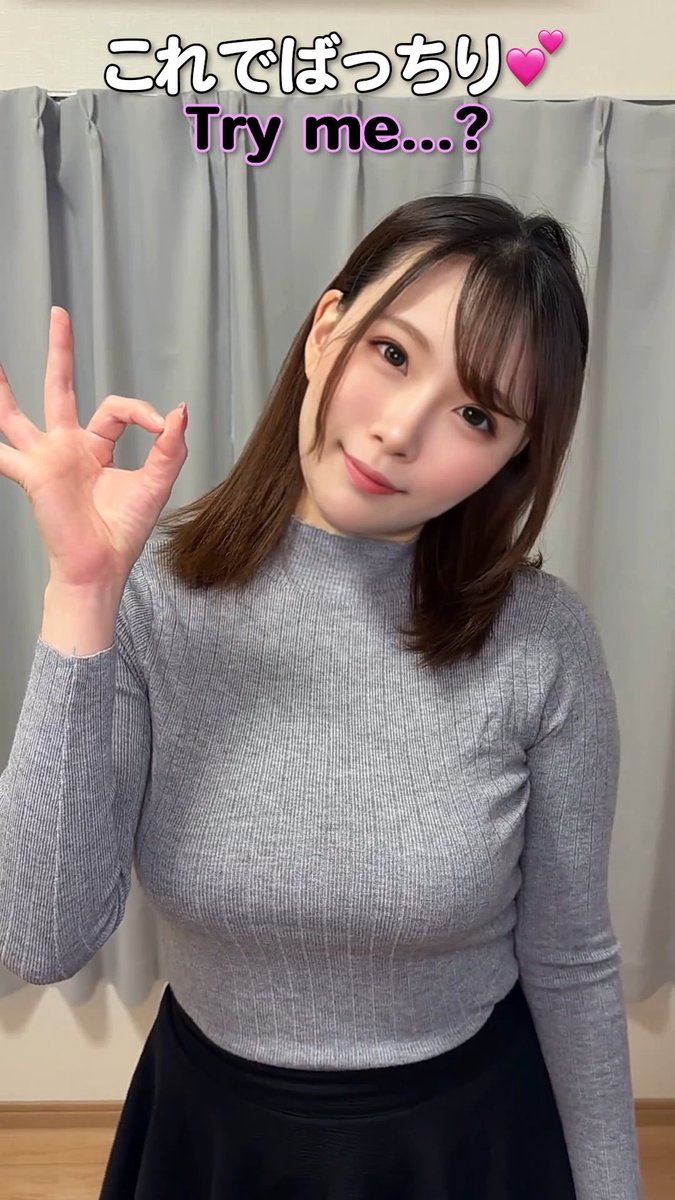 A young woman with brown hair poses in a gray ribbed turtleneck, making an "OK" hand gesture while smiling softly against a gray curtain backdrop. The text overlay reads "これでばっちり💕 Try me...?" which complements the caption's playful, suggestive tone about being "broken" by the gesture.