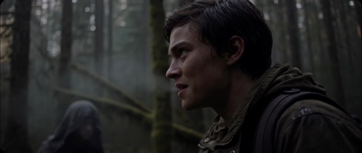 A still from a film showing a young man with a concerned expression in a misty forest, wearing a green jacket and backpack, with another figure visible in the background.