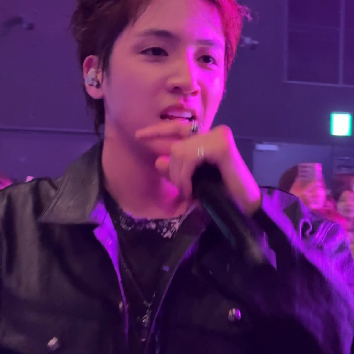A young male performer sings into a microphone on a stage illuminated with purple and pink lighting, wearing a black leather jacket and an earpiece.