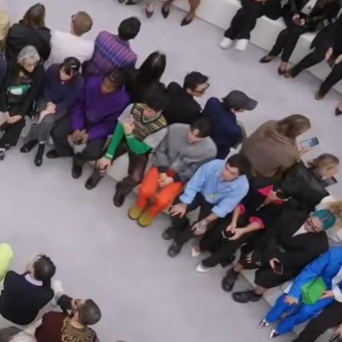 An overhead shot shows a crowd of people seated in rows at what appears to be a fashion show or event, dressed in various stylish clothing.