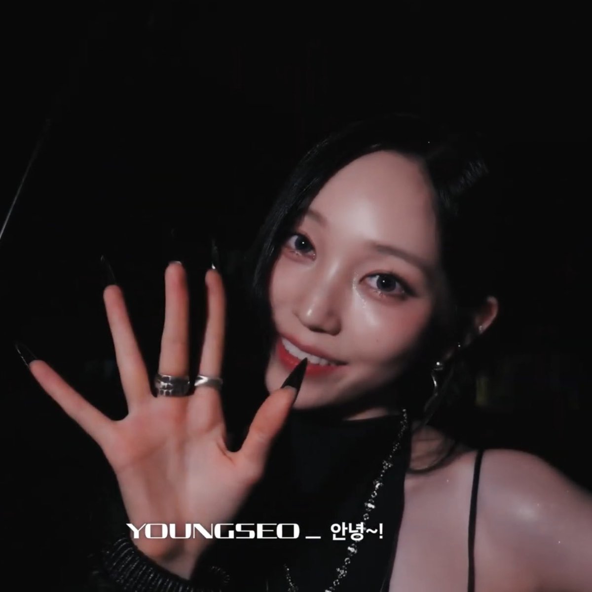 A dark close-up selfie of a smiling young woman holding up her hand with long black nails and rings, with text 'YOUNGSEO _ 안녕~!' overlaid.