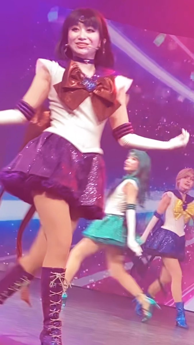 Performers on stage dressed in Sailor Moon-inspired costumes; the woman in the foreground sports a white top, large red bow, and purple skirt.