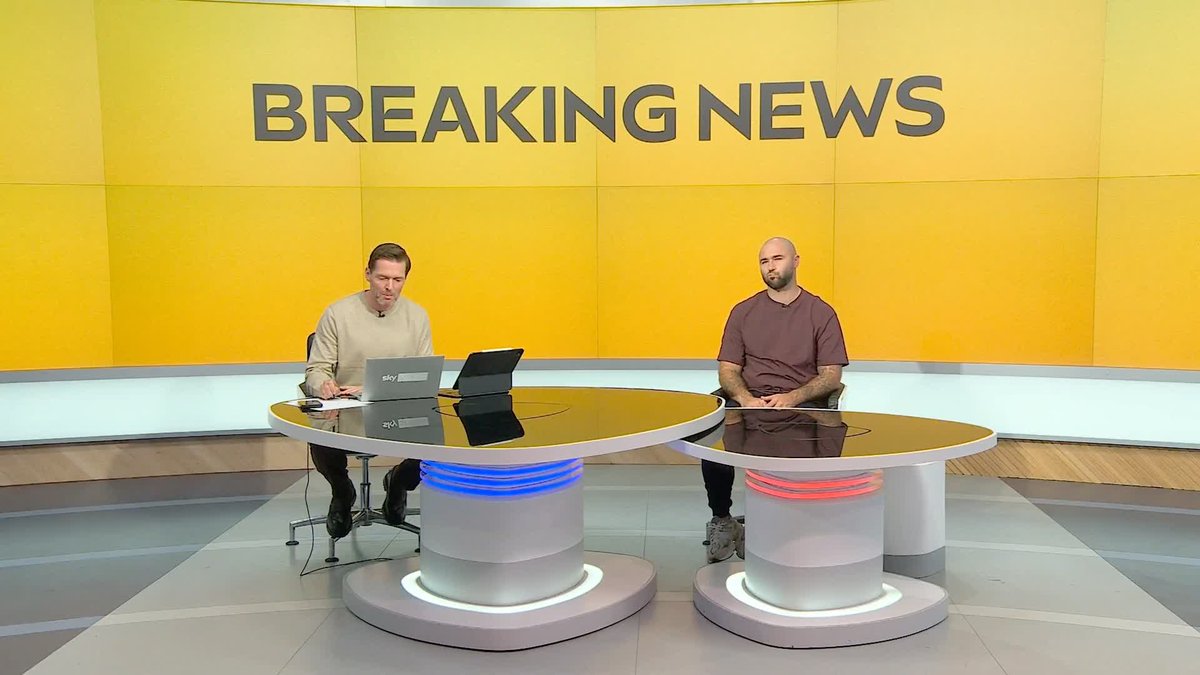 Two news anchors sitting at separate circular desks with glowing bases, in front of a bright yellow backdrop displaying the words 'BREAKING NEWS'.