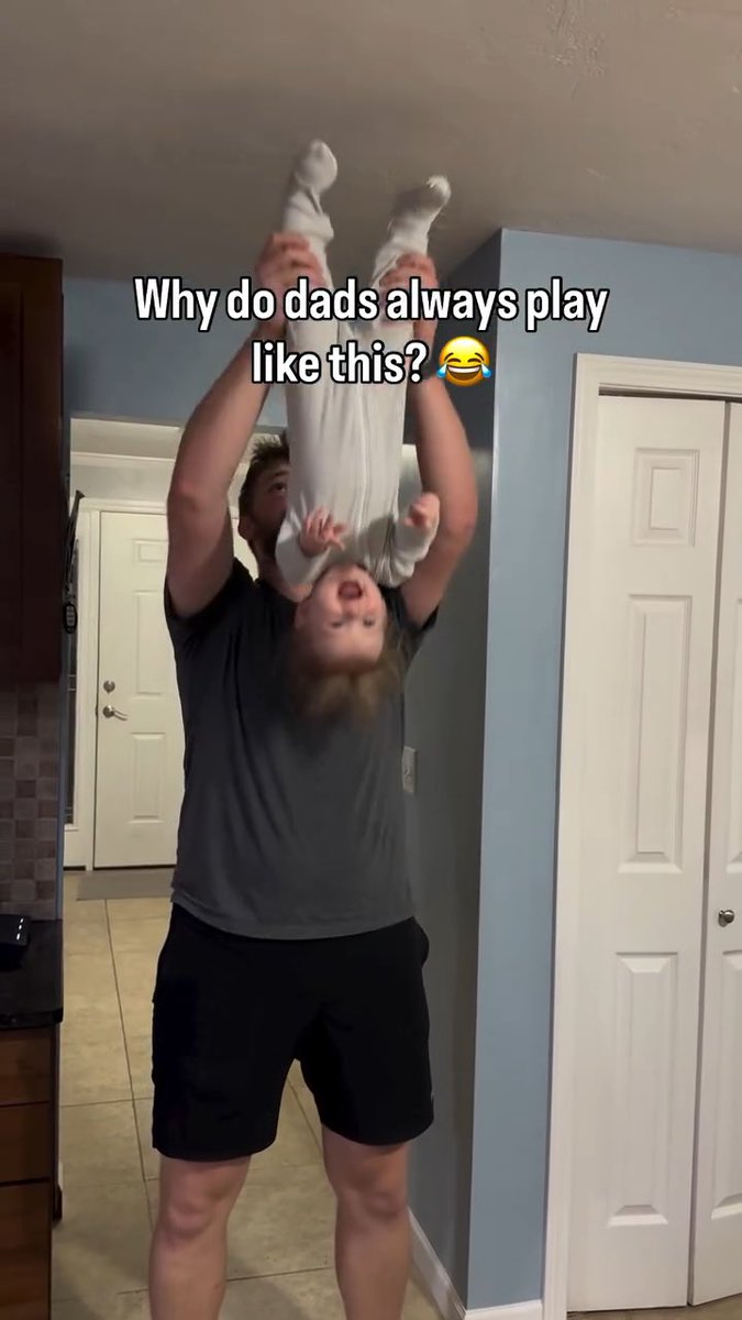 A father in a black shirt and shorts stands in a home hallway, playfully holding his laughing baby upside down, with text reading 'Why do dads always play like this?'