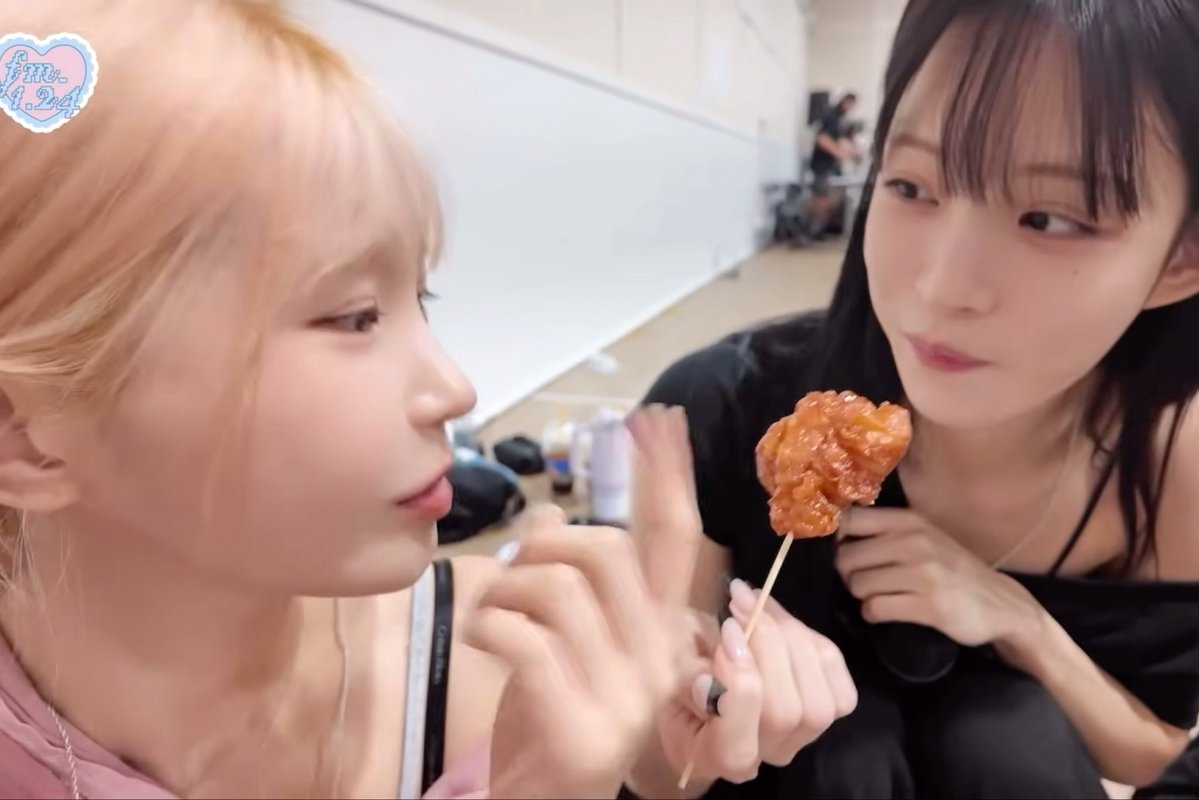 Two young women in a backstage setting; one with blonde hair looks at a piece of fried food on a stick held by the other woman with dark hair, who gazes at the food.