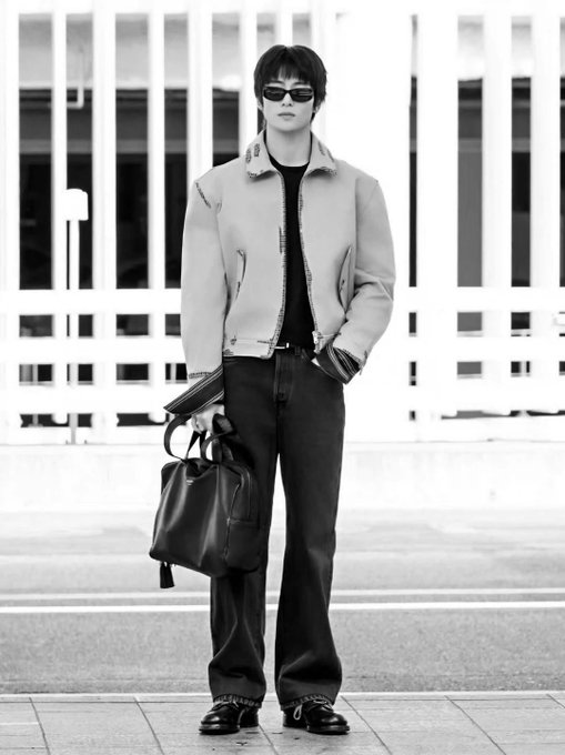 A black and white fashion photo of a person wearing dark sunglasses, a light-colored sherpa jacket, dark pants, and chunky shoes, holding a large black leather bag.