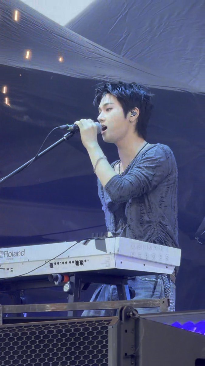 Concert photograph of a young male Asian singer with dark hair performing at a Roland keyboard, singing into a microphone on stage with dramatic lighting.