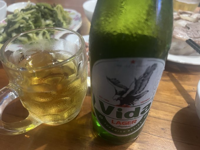A glass mug filled with a golden beverage sits next to a green bottle of 'Vida' Premium Lager on a dining surface.