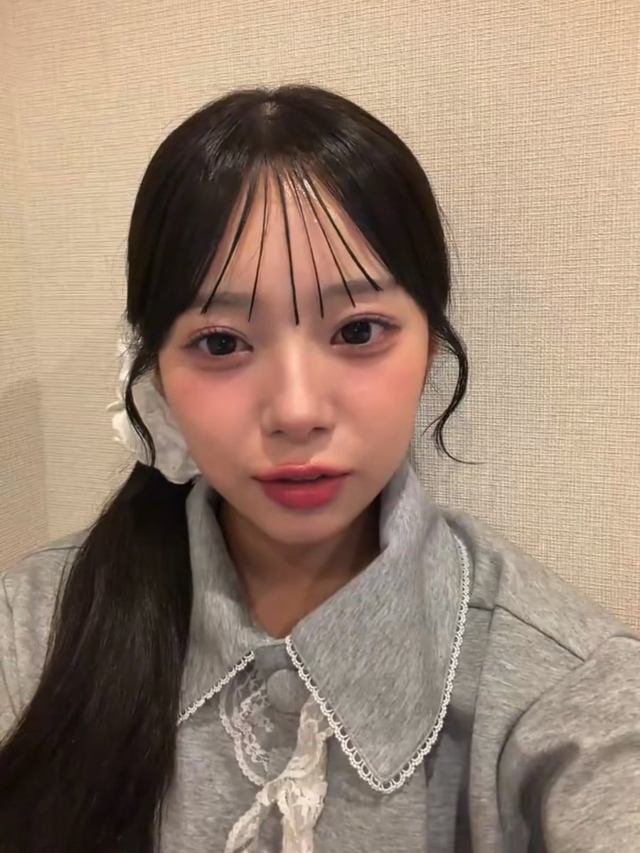 A close-up portrait of a young woman with dark hair, blunt fringe, heavy blush, and red lipstick, wearing a gray top with white lace details.