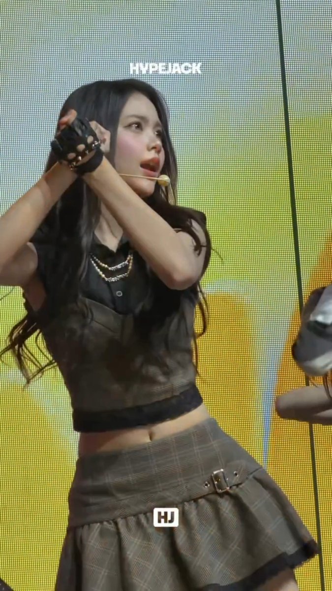 A performer with long dark hair on stage wearing a black crop top, plaid skirt, and studded gloves, performing with a microphone against a bright yellow background, branded 'HYPEJACK' and 'HJ'.