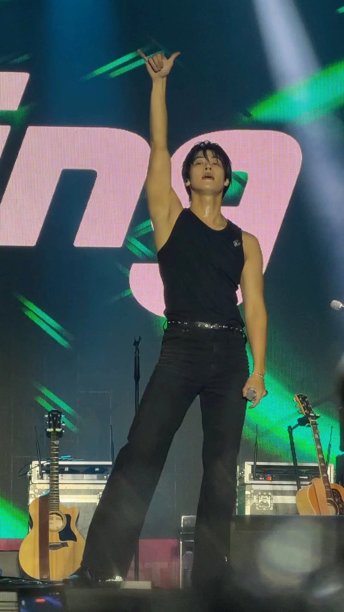 A male performer in a black tank top and black jeans points upward with one hand while holding a microphone, with acoustic guitars and a green-lit backdrop visible on stage.