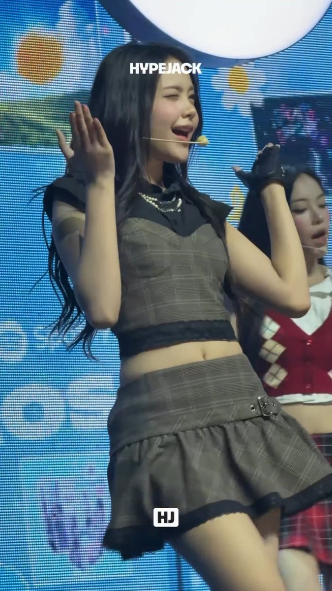 A young woman with long dark hair performs on stage wearing a plaid two-piece outfit with a black collar and fingerless gloves, with 'HYPEJACK' and 'HJ' watermarks visible.