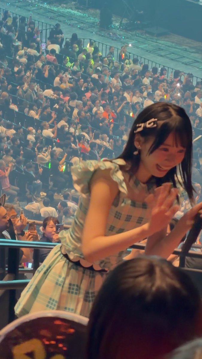 A young woman with dark hair and a hair clip wearing a pastel plaid dress claps enthusiastically at a concert, with a large crowd of fans holding glow sticks visible in the background.