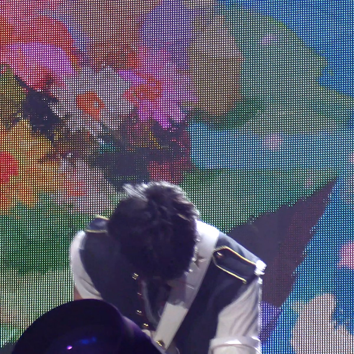 A person with dark hair bows their head while performing on stage, wearing a white shirt with gold-trimmed dark epaulets against a colorful LED screen background.