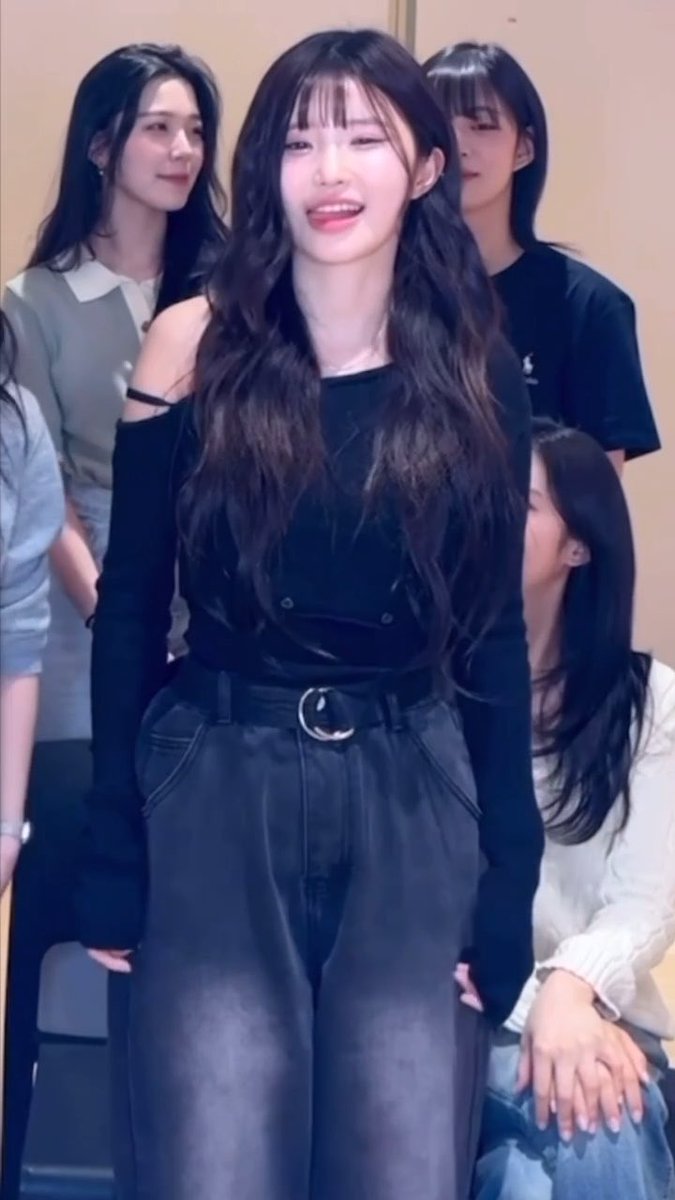 A young woman with long dark wavy hair wearing a black off-the-shoulder top and dark jeans smiles at the viewer, surrounded by other young women.