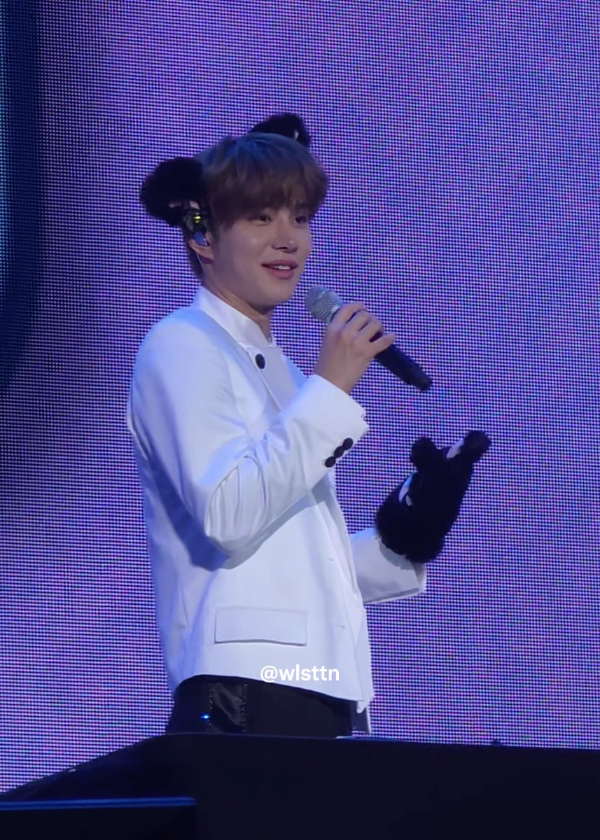 A young performer in a white jacket holds a microphone and smiles on stage, with a watermark reading '@wlstttn' visible.