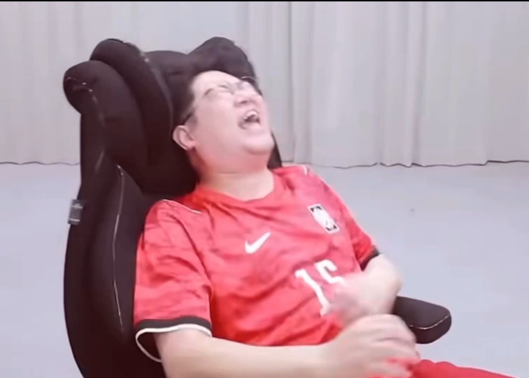 A young man in a red soccer jersey (#15) laughing hysterically while sitting in a black office chair.