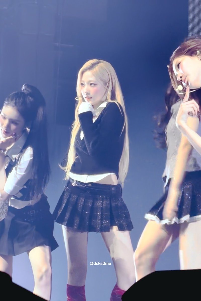 A photograph of K-pop idol Karina from aespa performing on stage in a black sweater over a white collared shirt and a black pleated skirt, with a '@dsks2me' watermark.