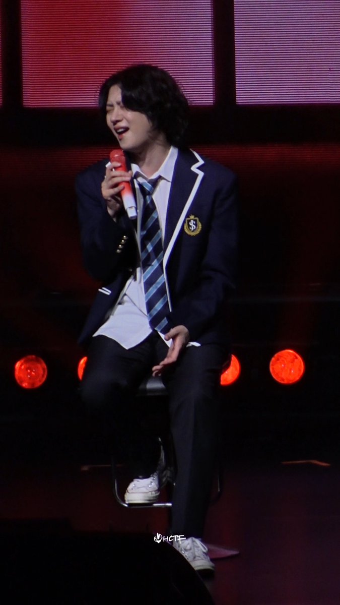 A young male performer in a navy school uniform blazer sings into a red microphone while seated on stage with red lights in the background.