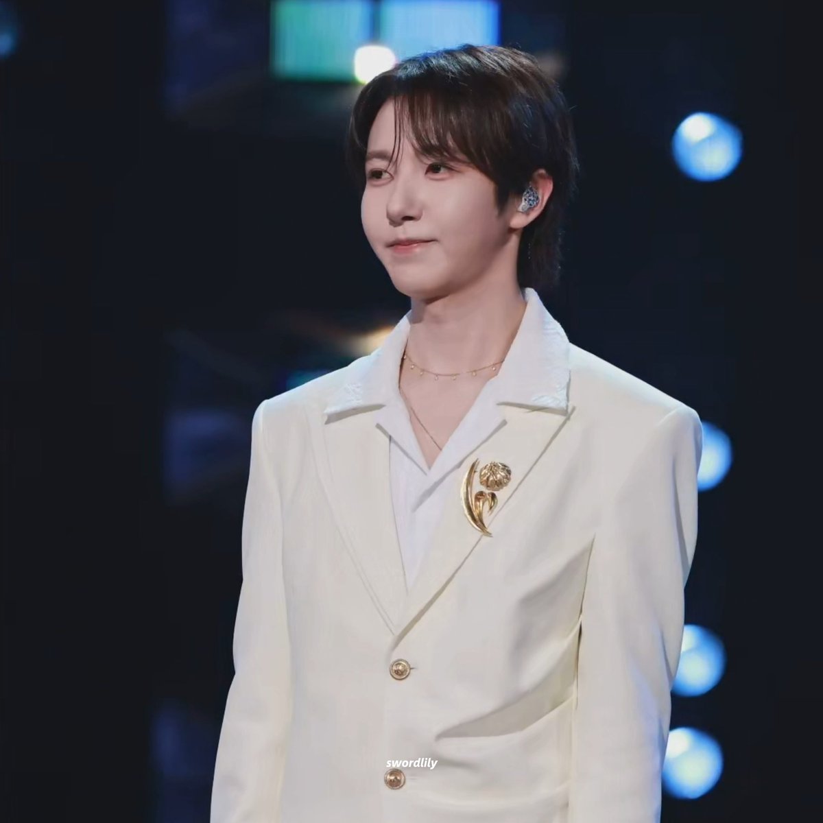 A young man with dark hair, wearing a white blazer with a gold pin, stands on a stage with the watermark 'swordlily' visible at the bottom.