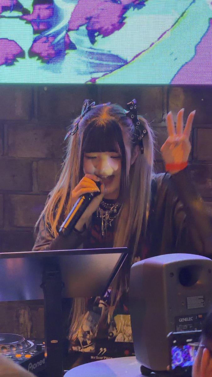 A woman with long hair and decorative clips sings into a microphone while raising her hand, performing behind Pioneer DJ equipment with a Genelec speaker, set against a brick wall with a colorful projection screen above.
