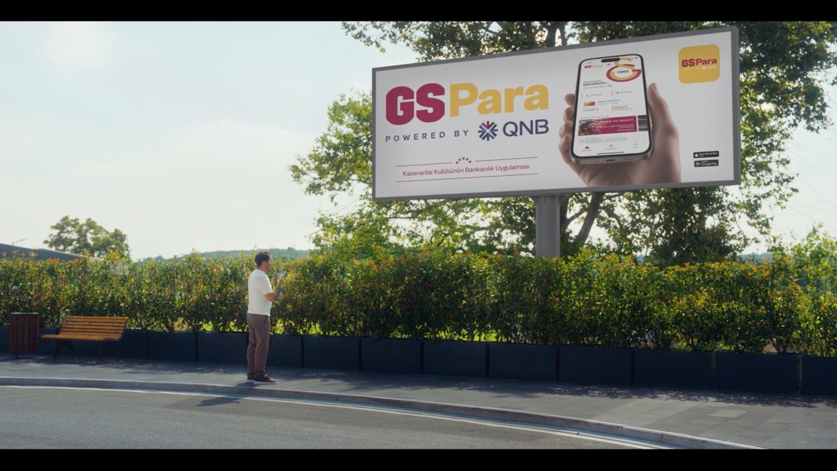 A man in a white shirt and brown pants stands on a roadside looking at his phone beneath a large billboard advertising "GS Para Powered by QNB," a banking app for fans of the Turkish sports club Galatasaray, with the Turkish text "Kazananlar Kulübünün Bankacılık Uygulaması" below. The image humorously illustrates the post's joke about unnamed sponsors pulling out, since GS Para (Galatasaray's financial partner) appears as a lone sponsor rather than a typical lineup of multiple advertisers.