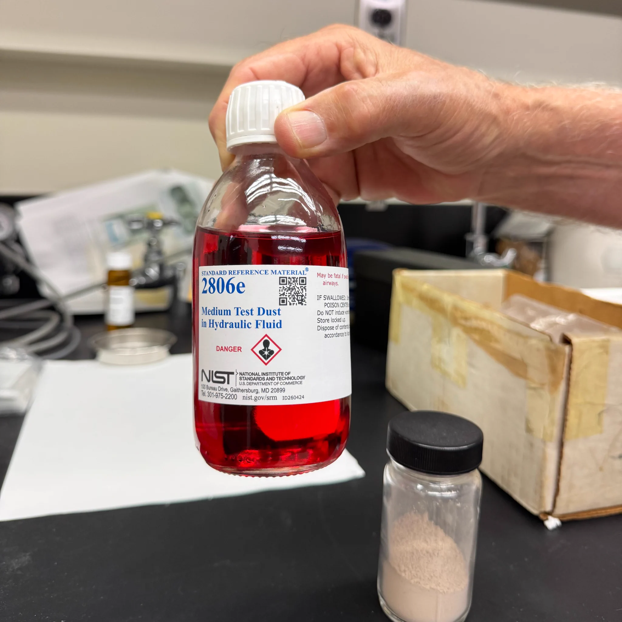 A hand is holding a clear bottle labeled "NIST 2806e Medium Test Dust in Hydraulic Fluid," filled with a red liquid. The bottle is in a laboratory setting, with various equipment visible in the background.