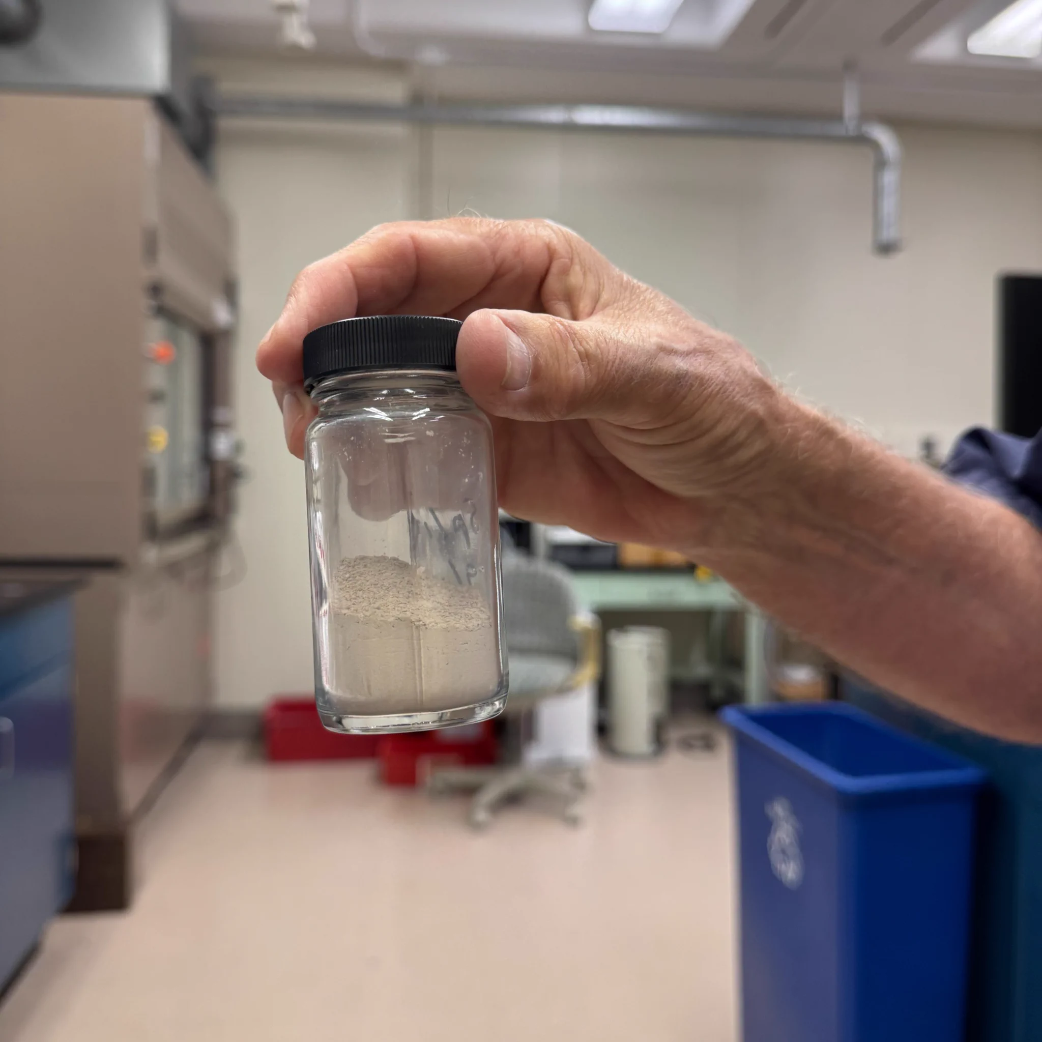 A hand is holding a small glass jar with a black lid containing a light-colored powder. The background shows a laboratory setting with equipment and furniture.