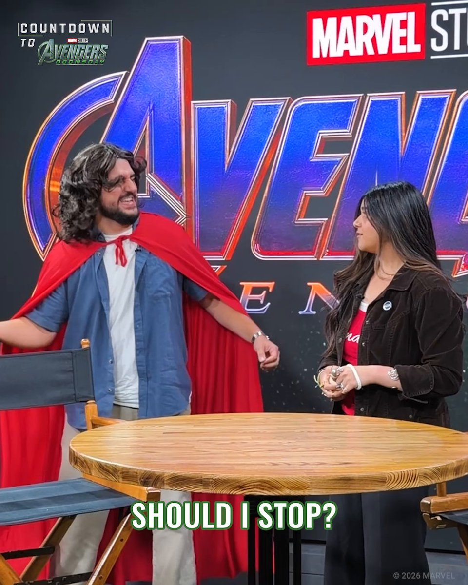 A man wearing a red cape stands in front of an "Avengers Doomsday" backdrop, looking toward a woman across a wooden table, with the caption "SHOULD I STOP?" appearing at the bottom in green text. The image ties to the soap-on-a-rope concept by showing the man with a rope/cape hanging down like a lanyard, prompting the humorous question of whether he should stop swinging it during the promotional appearance.
