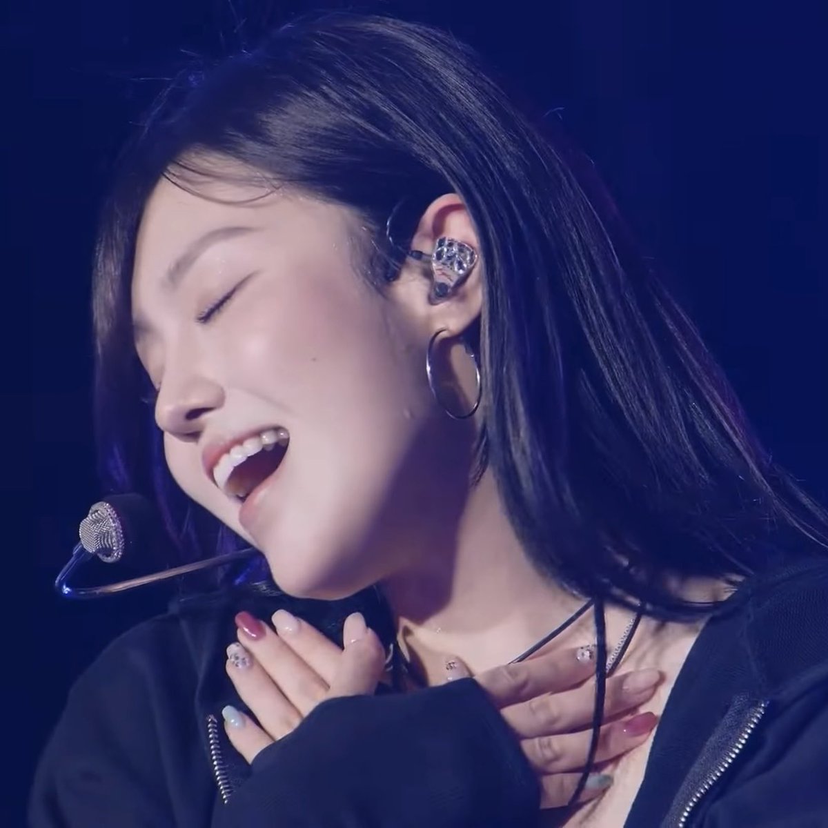 A close-up shot captures a young woman singing passionately into a microphone on stage, wearing a black jacket, hoop earrings, and custom in-ear monitors.