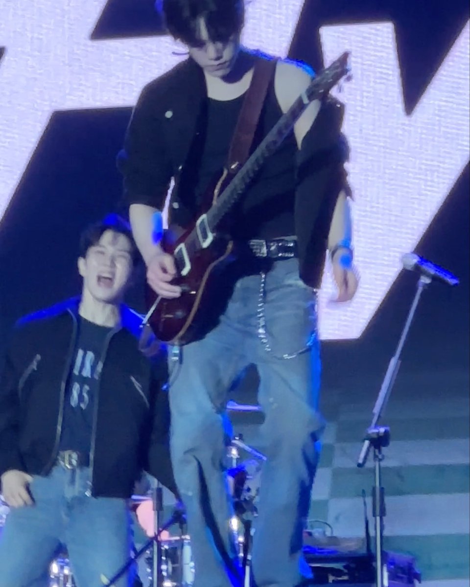Two performers are on stage; one in the background plays an electric guitar and stands elevated, wearing a black jacket and jeans, while another smiles in the foreground.