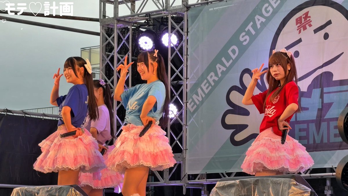 Three young female performers on the 'EMERALD STAGE' pose with peace signs while dressed in brightly colored tops and matching tiered pink ruffled skirts, holding microphones on an outdoor stage.