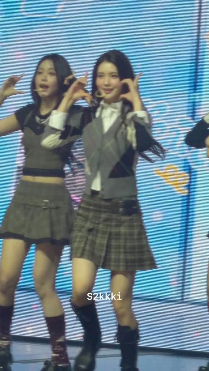 Two young women perform on stage in matching plaid schoolgirl-style outfits with skirts and ties, posing with hand gestures (watermarked S2kkki).