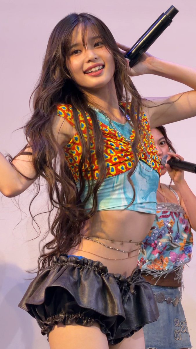 A smiling young woman with long wavy hair performs on stage, wearing a colorful patterned crop top over a turquoise shirt and a black ruffled skirt, holding a microphone.