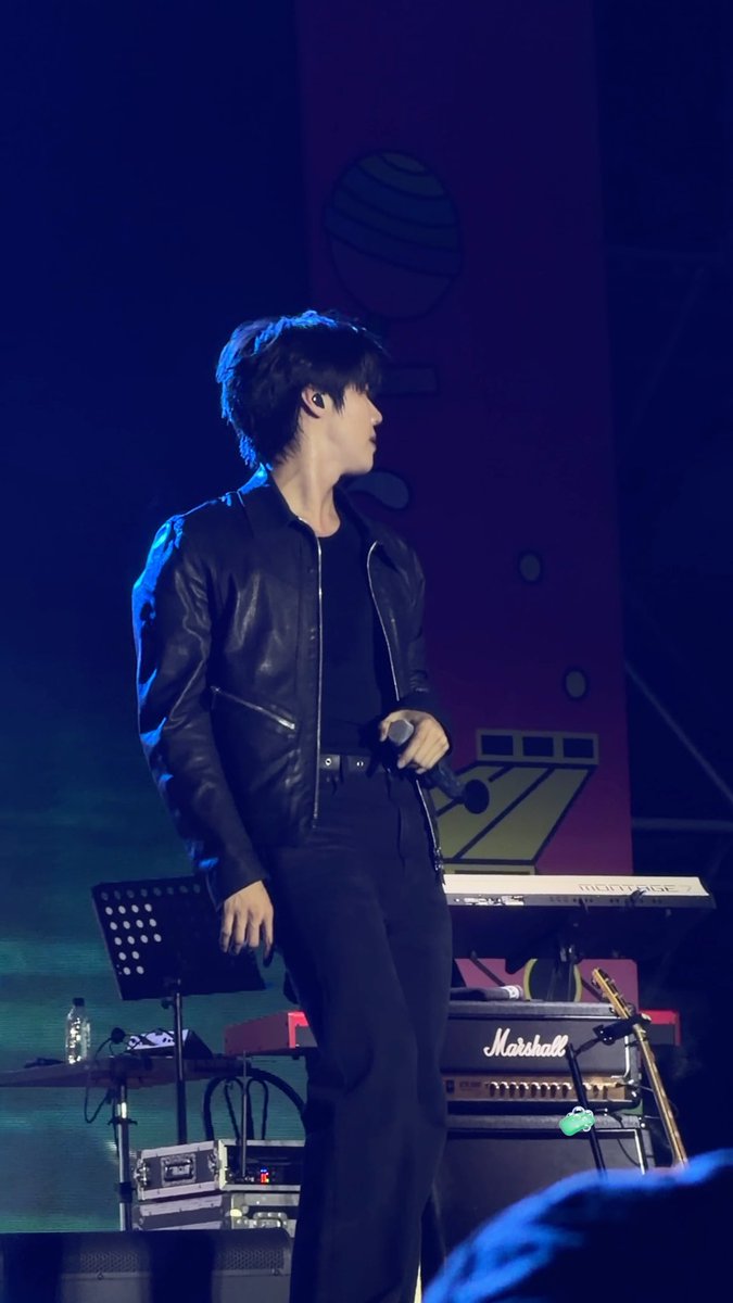 A man in a black leather jacket stands on a concert stage holding a microphone, with a keyboard marked 'MONTAGE' and a Marshall amplifier visible behind him, along with blue stage lighting.