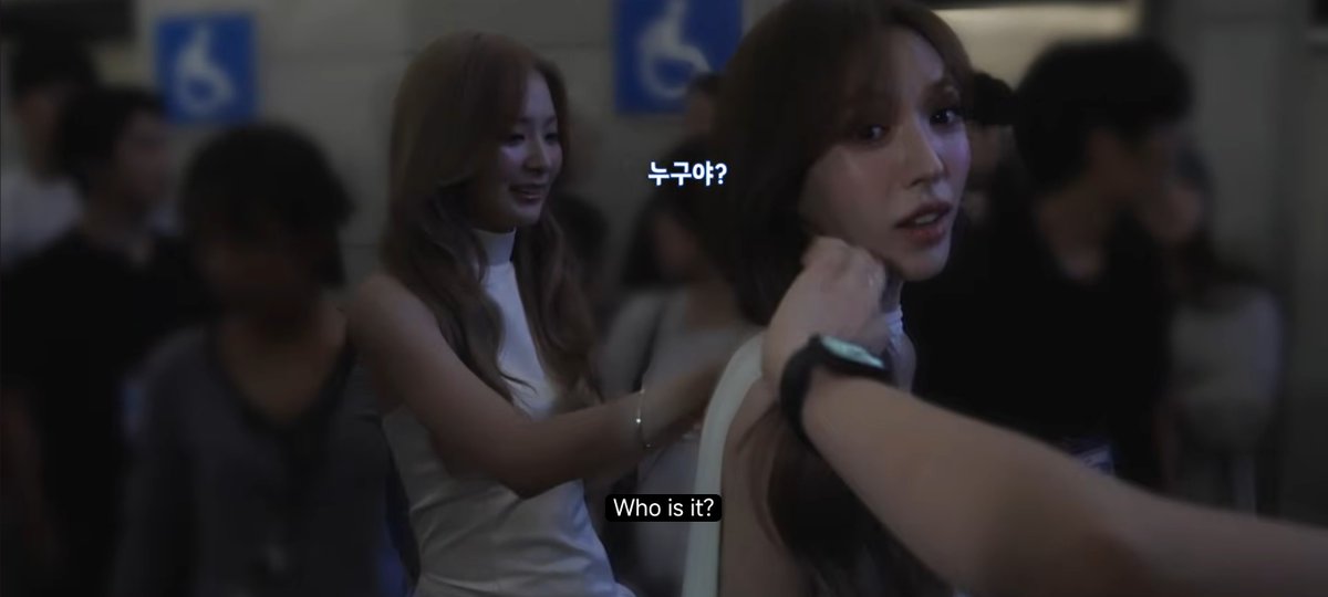 A scene from a drama showing two women in a crowded setting, with subtitles 'Who is it?' in English and '누구야?' in Korean.