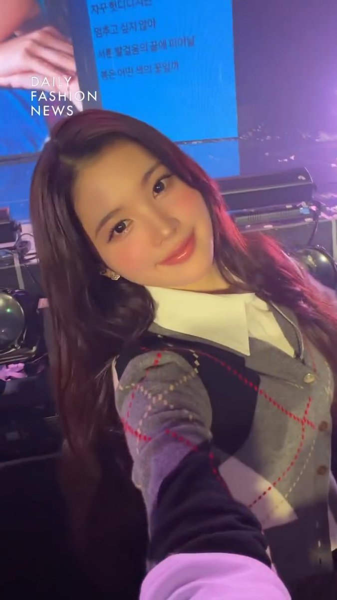 A selfie of a smiling young woman with long dark hair wearing a gray argyle sweater, with 'DAILY FASHION NEWS' text overlaid and Korean text visible on a screen in the background.