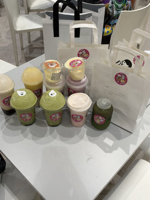 A table displays an assortment of colorful drinks in plastic cups with dome lids, surrounded by several white paper takeout bags featuring cartoon character logos.