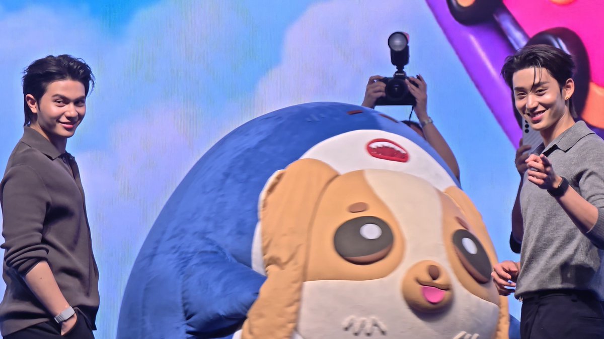 Two smiling young men stand on stage flanking a large, cute dog mascot wearing a blue outfit, while a photographer with a camera captures the moment from behind. The image relates to the post's hashtags "#WilliamEst" and "#Wesley" featuring "Happy Family" members at what appears to be the "GMMTV Fanival 2026" event, promoting their new "Happy Family" project.