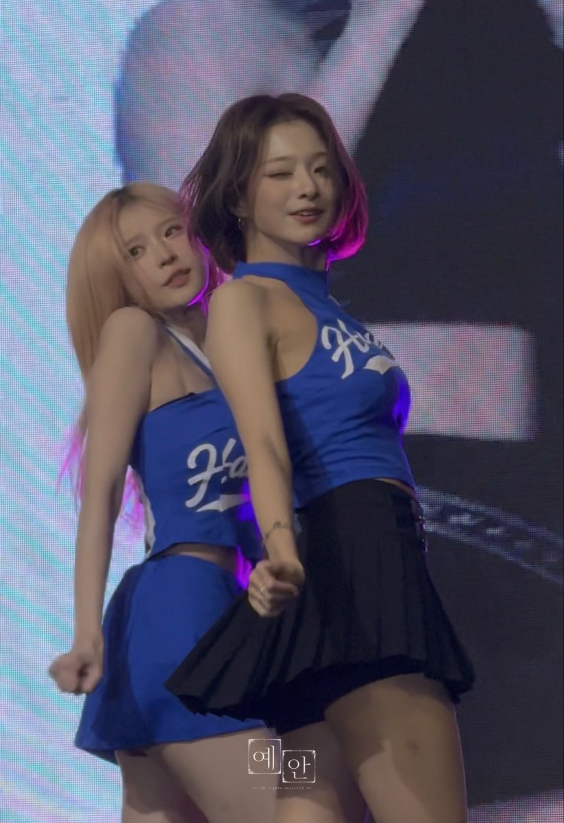 Two performers in matching blue 'Hun' cheerleader-style tops and black pleated skirts dance on stage, with a Korean watermark reading '예완' visible.