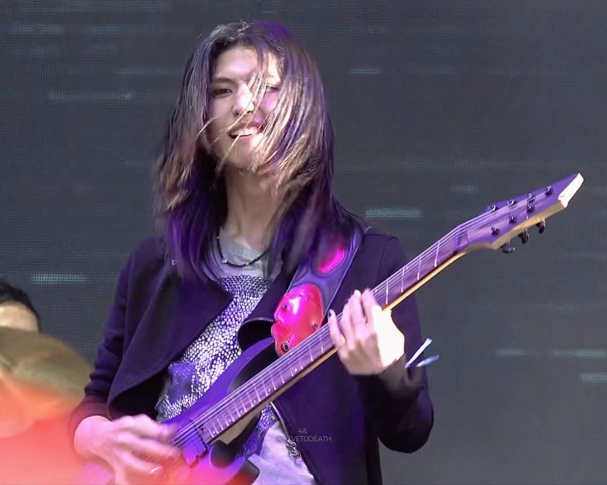 A guitarist with long hair performs on stage playing an electric guitar, wearing a dark jacket over a patterned shirt.