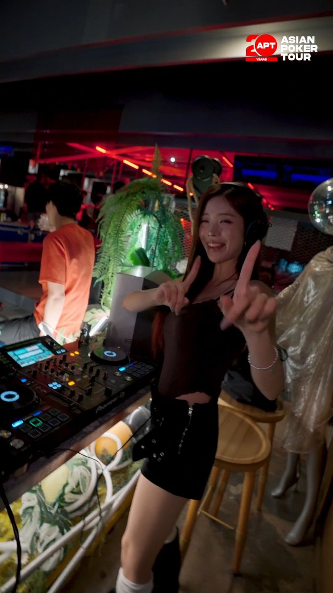 A young woman wearing headphones smiles and points at the camera while standing next to DJ equipment, with an 'APT ASIAN POKER TOUR' logo visible.